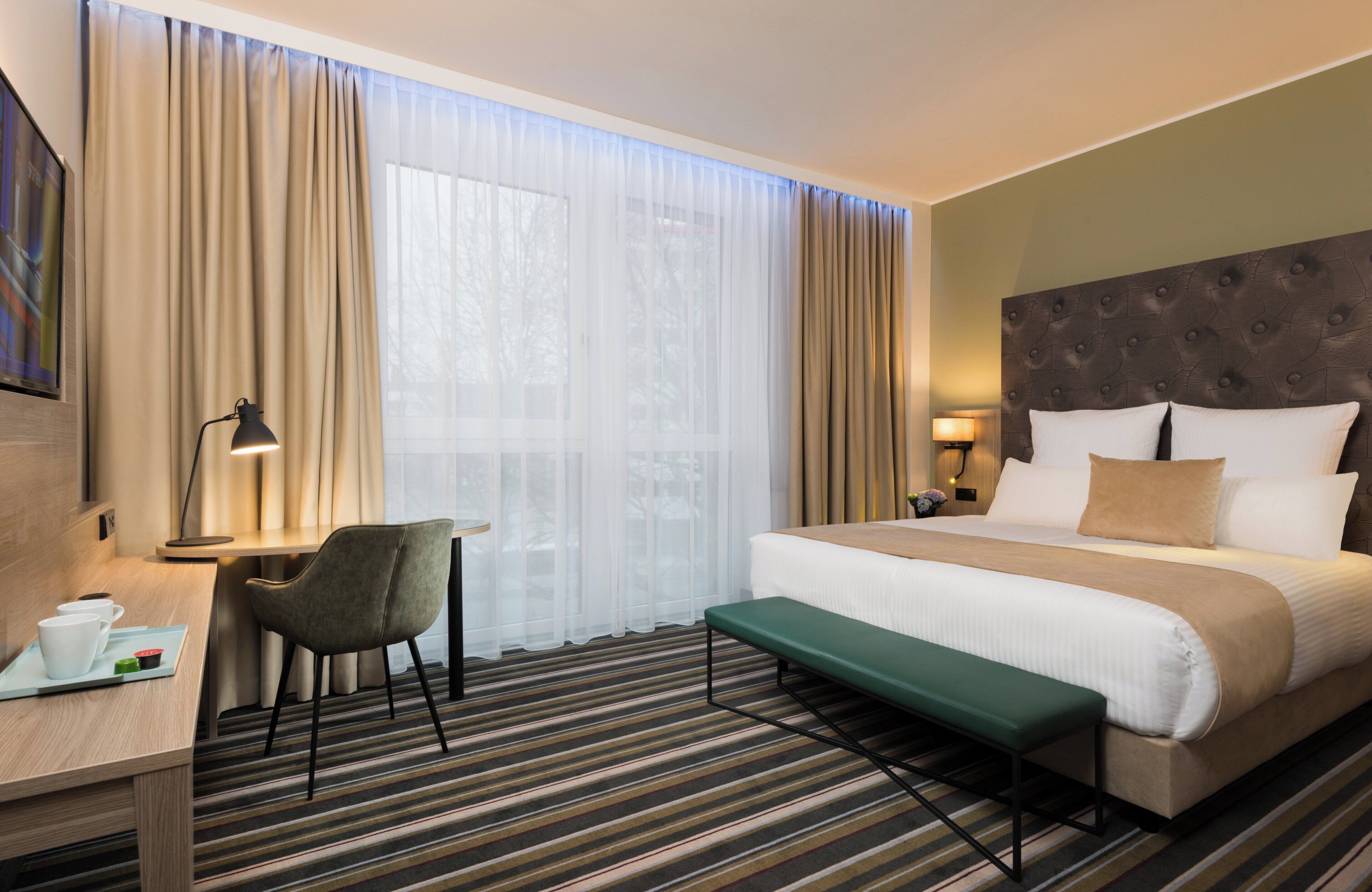 A hotel room with a bed, a desk, a chair, a bench, and a large window with curtains.