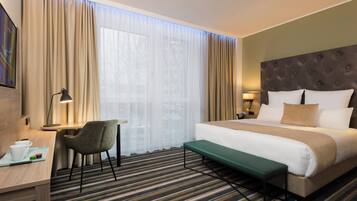 A hotel room with a bed, a desk, a chair, a bench, and a large window with curtains.