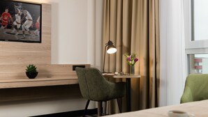 A hotel room with a wooden desk, a green chair, a small table with a lamp, a vase with flowers, and a television displaying a basketball game. - Leonardo Hotel Offenbach Frankfurt (Offenbach am Main)