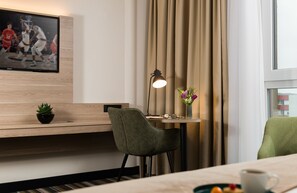 Habitación de hotel con un escritorio de madera, una silla verde, una mesita con lámpara, un jarrón con flores y un televisor mostrando un partido de baloncesto.