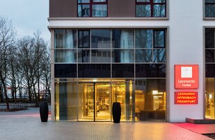 A modern hotel entrance with large glass doors, a sign indicating it's the Leonardo Hotel in Frankfurt, and a well-lit interior visible through the doors.