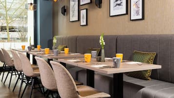 A restaurant with a long table, cushioned chairs, and framed artwork on the wall.