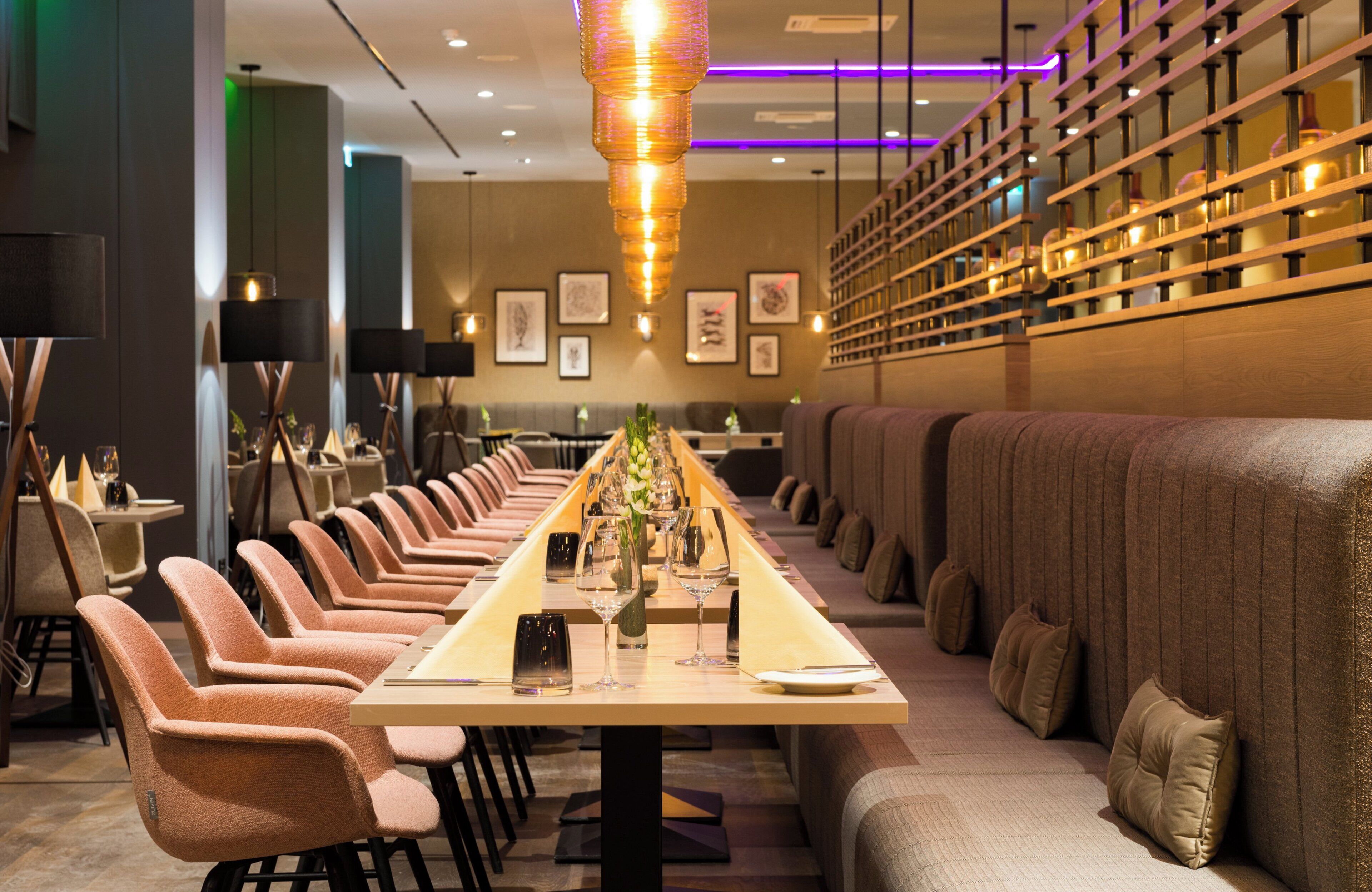 A restaurant with a long communal table, cushioned seating, and pendant lighting.
