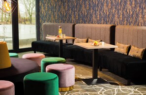 A modern lounge area with a sofa, colorful ottomans, a table with a bottle and glasses, and a patterned carpet. - Leonardo Hotel Offenbach Frankfurt (Offenbach am Main)