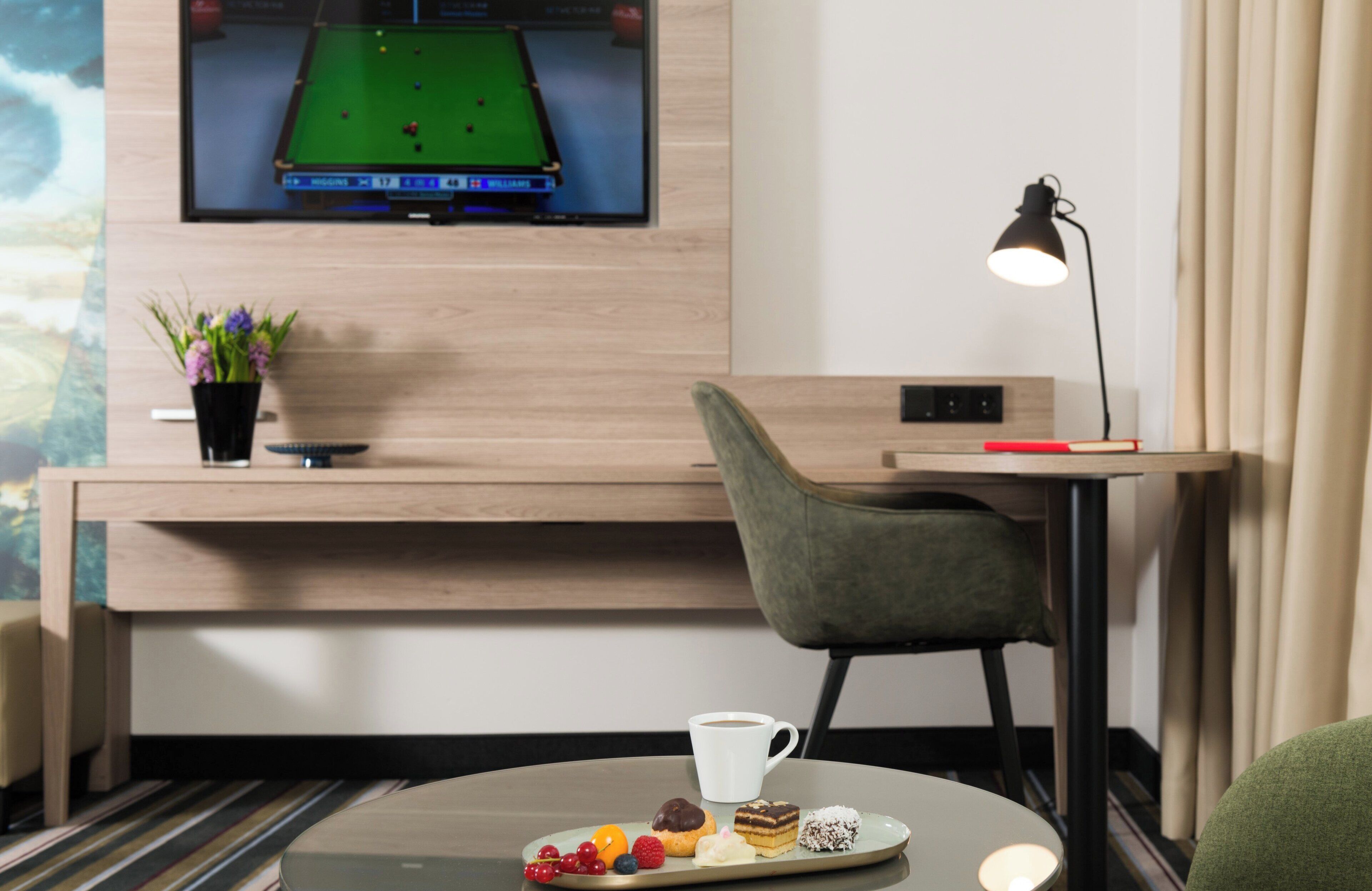 Una habitación de hotel moderna con mesa de billar, un televisor mostrando un partido de billar, un escritorio con lámpara y una pequeña mesa con aperitivos.