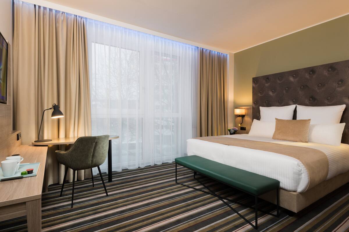 A hotel room with a bed, a desk with a chair, a bench, a lamp, and a window with curtains.