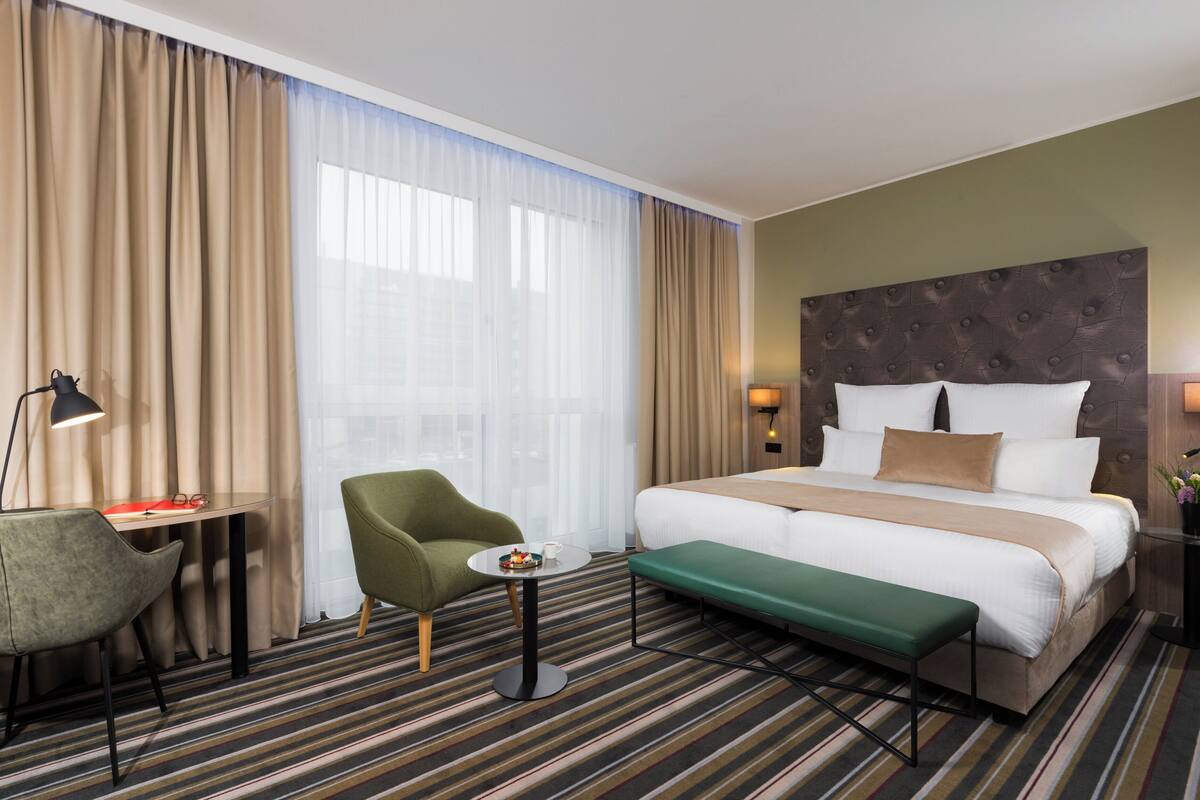 A hotel room with a large bed, a green armchair, a small table with a lamp, and a bench.