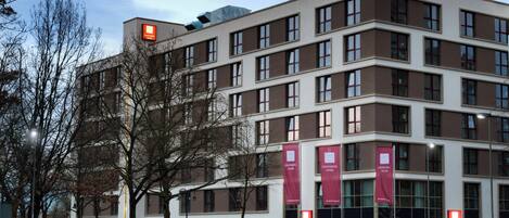 Un moderno edificio de hotel con varios pisos, grandes ventanales y pañoletas rojas que indican el nombre del hotel.