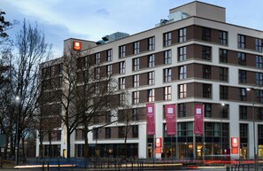 Un moderno edificio de hotel con varios pisos, grandes ventanales y pañoletas rojas que indican el nombre del hotel.
