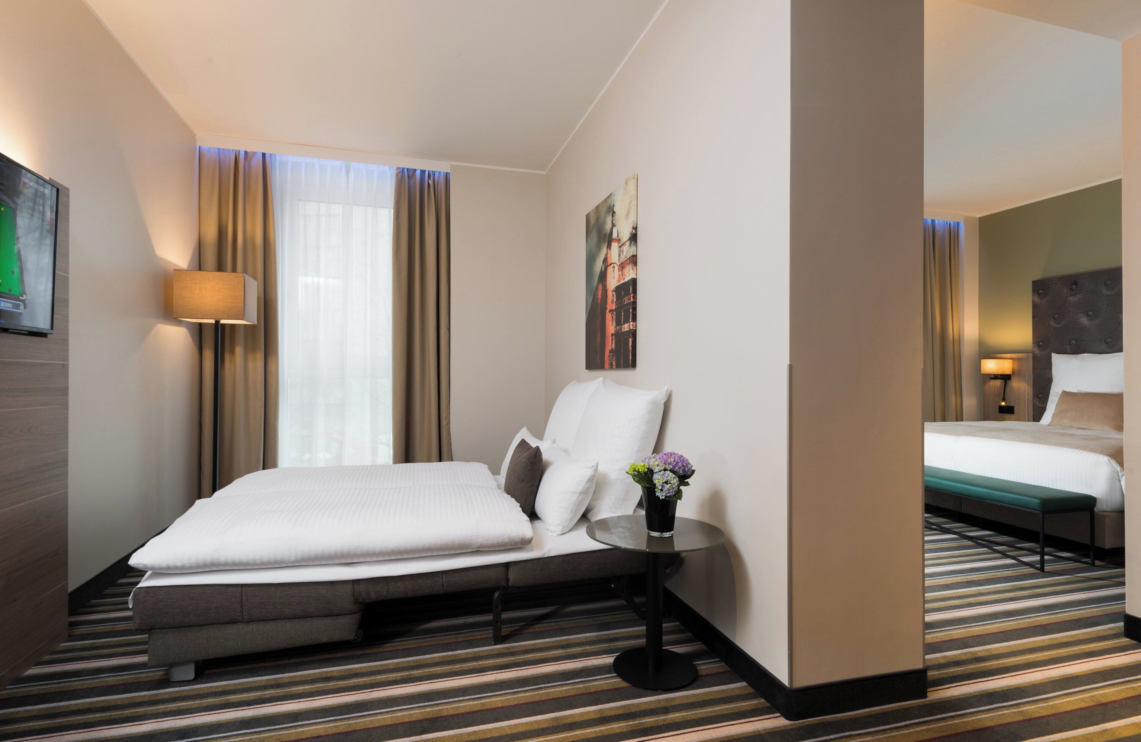 Ein Hotelzimmer mit Bett, Nachttisch, Lampe, einem Fenster mit Vorhängen und einem Bild an der Wand.