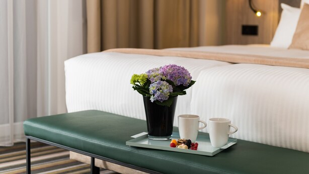 A hotel room with a neatly made bed, a bedside table with a tray of snacks and a cup, and a vase of flowers. - Leonardo Hotel Offenbach Frankfurt (Offenbach am Main)