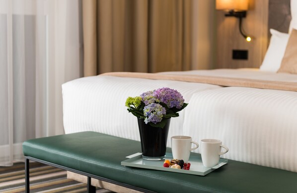 Una habitación de hotel con una cama bien hecha, una mesita de noche con una bandeja de aperitivos y una taza, y un jarrón con flores.