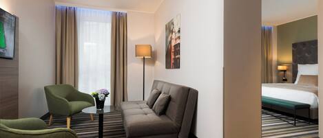 A hotel room with a sofa, armchair, a small table with a vase, a bed with a bench, and a painting on the wall.