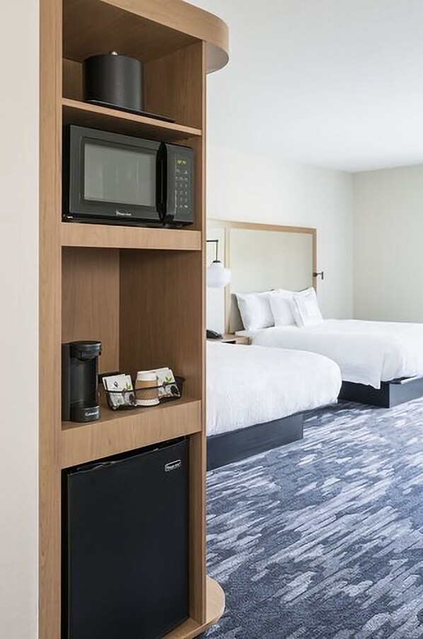 Habitación de hotel con microondas, cafetera y mini refrigerador en un estante, dos camas con sábanas blancas y alfombra gris.