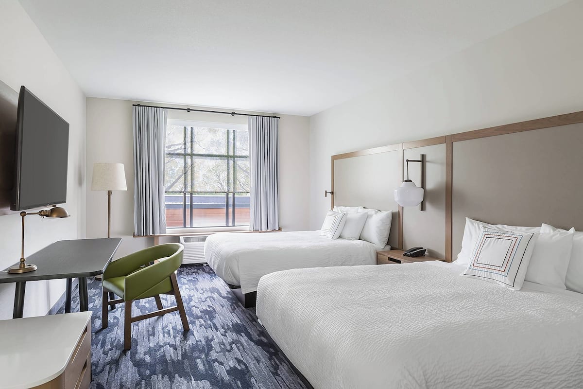 Ein Hotelzimmer mit zwei Betten, einem Schreibtisch, einem Stuhl, einem Fernseher und einem Fenster mit Vorhängen.