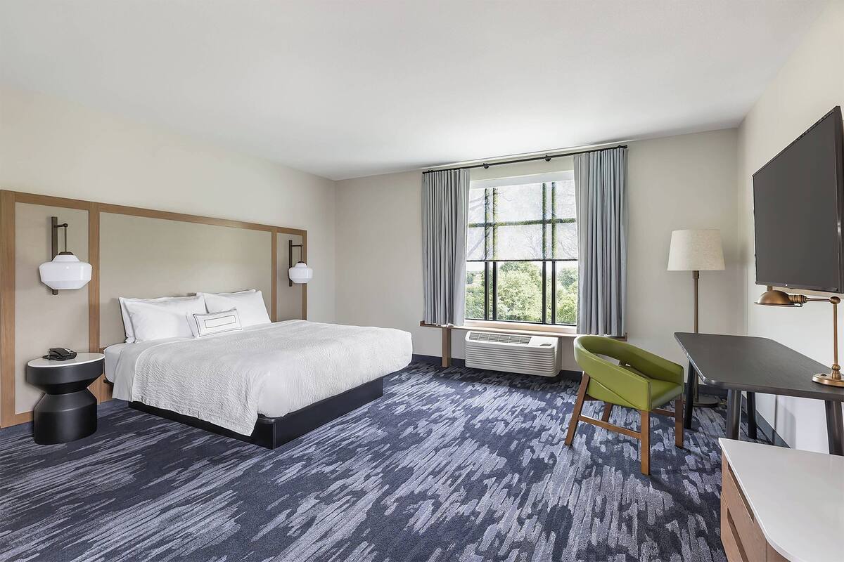 Ein Hotelzimmer mit einem großen Bett, einem Schreibtisch, einem Sessel, einem Fernseher und einem Fenster mit Vorhängen.