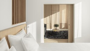 Un dormitorio con cama, almohadas, cabecera, ventana y vistas a otra habitación con planta y mesa.