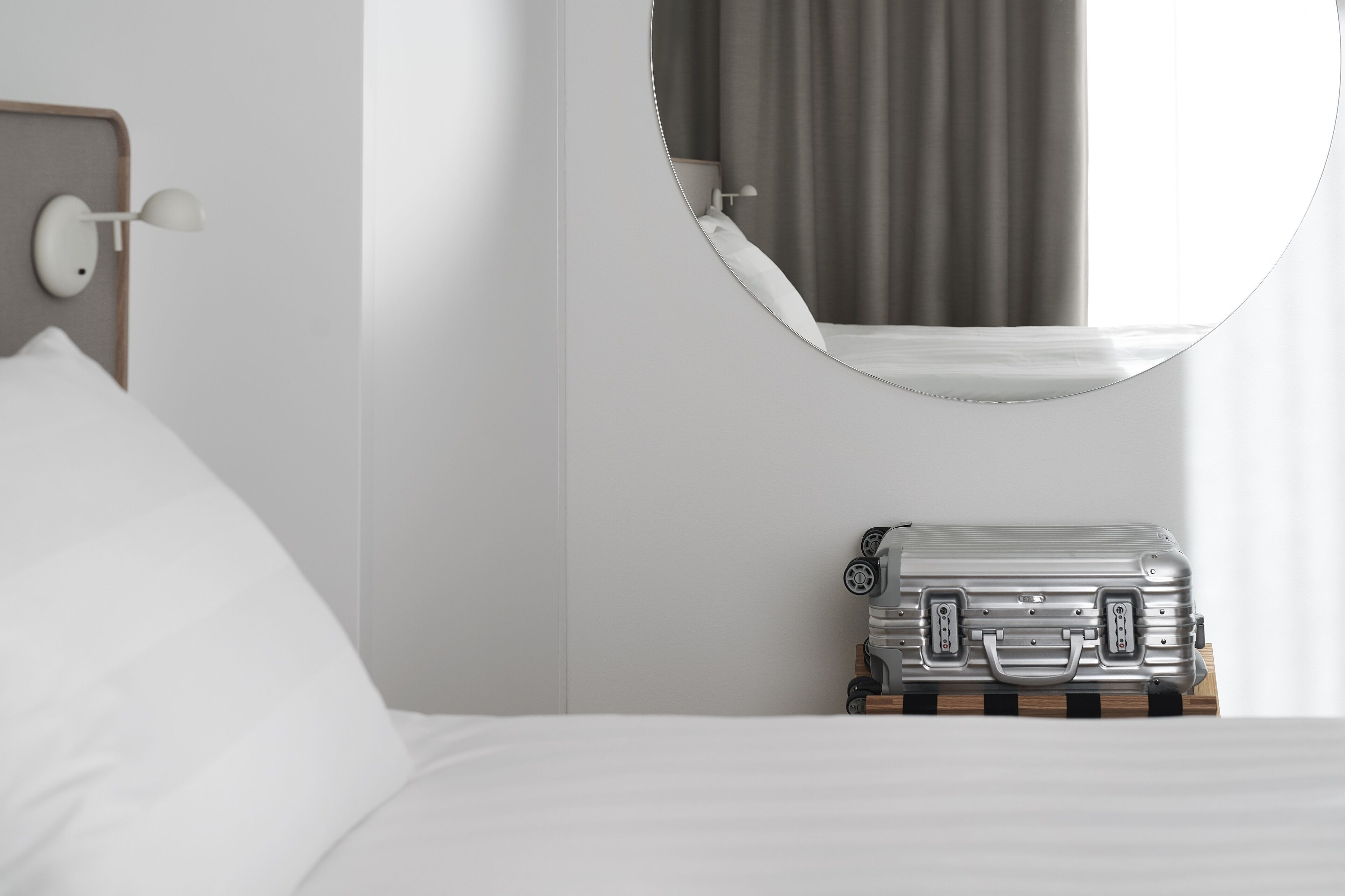 Una habitación de hotel con una cama, un espejo redondo, una lámpara de noche y una maleta sobre una mesita de madera.