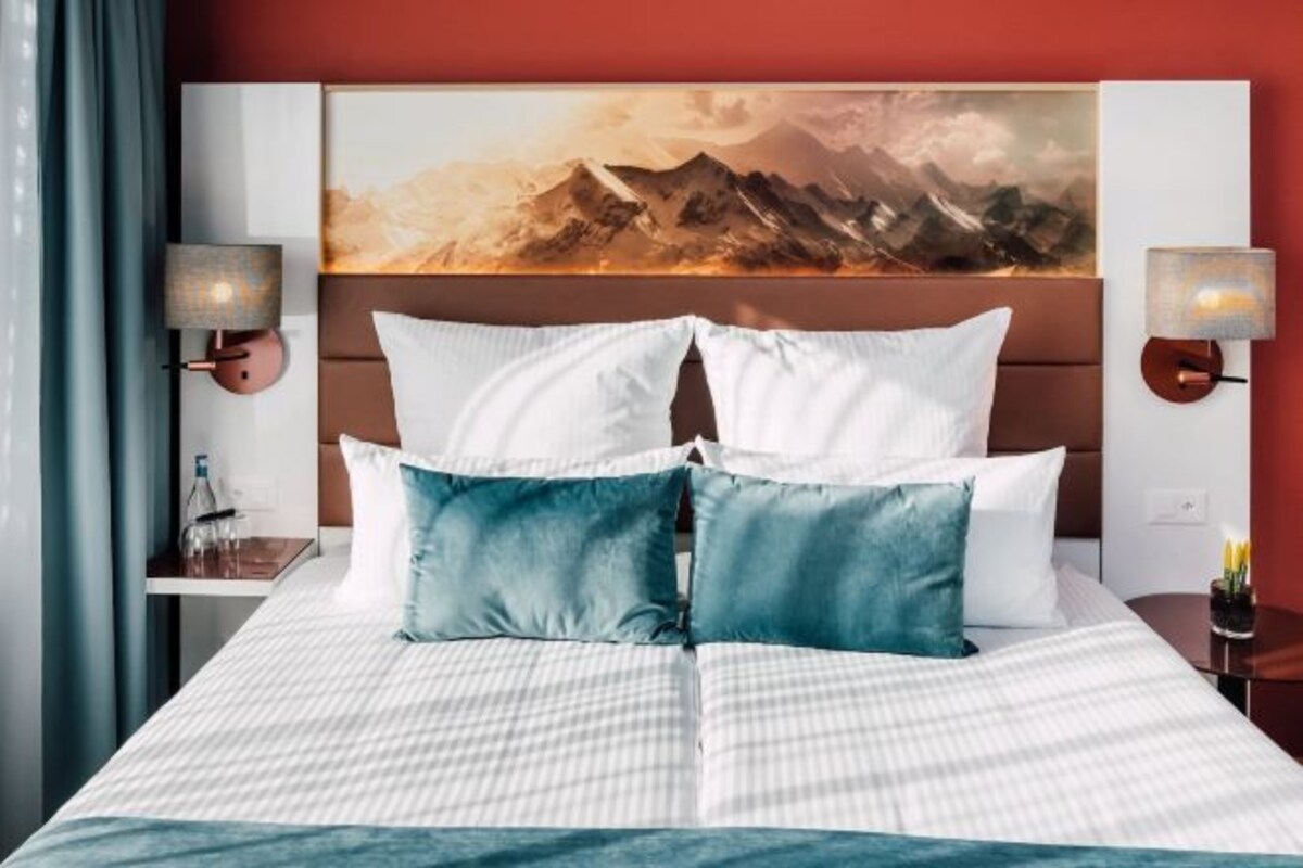 Een hotelkamer met een bed, een hoofdeinde met een schilderij, twee bedlampen en een nachtkastje met een fles.