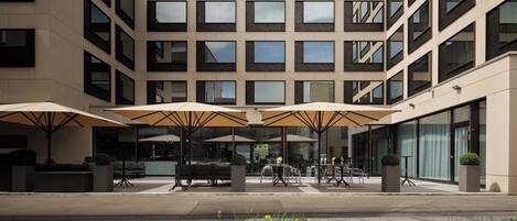 A modern building with multiple floors, outdoor seating with umbrellas, and a well-maintained garden area.
