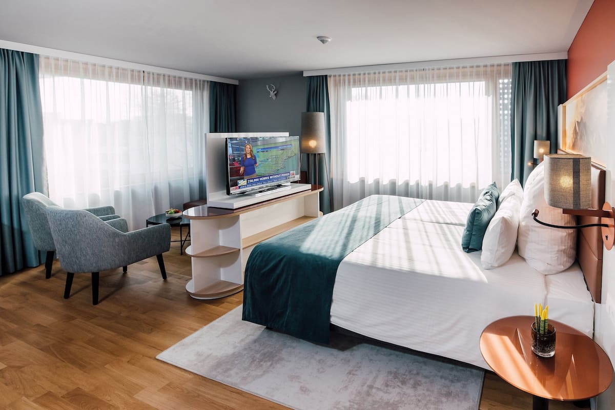 Une chambre d’hôtel moderne avec un grand lit, un bureau avec une télévision, une chaise et une petite table agrémentée d’une plante.