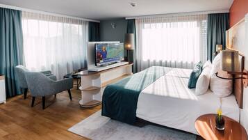 A modern hotel room with a large bed, a desk with a TV, a chair, and a small table with a plant.
