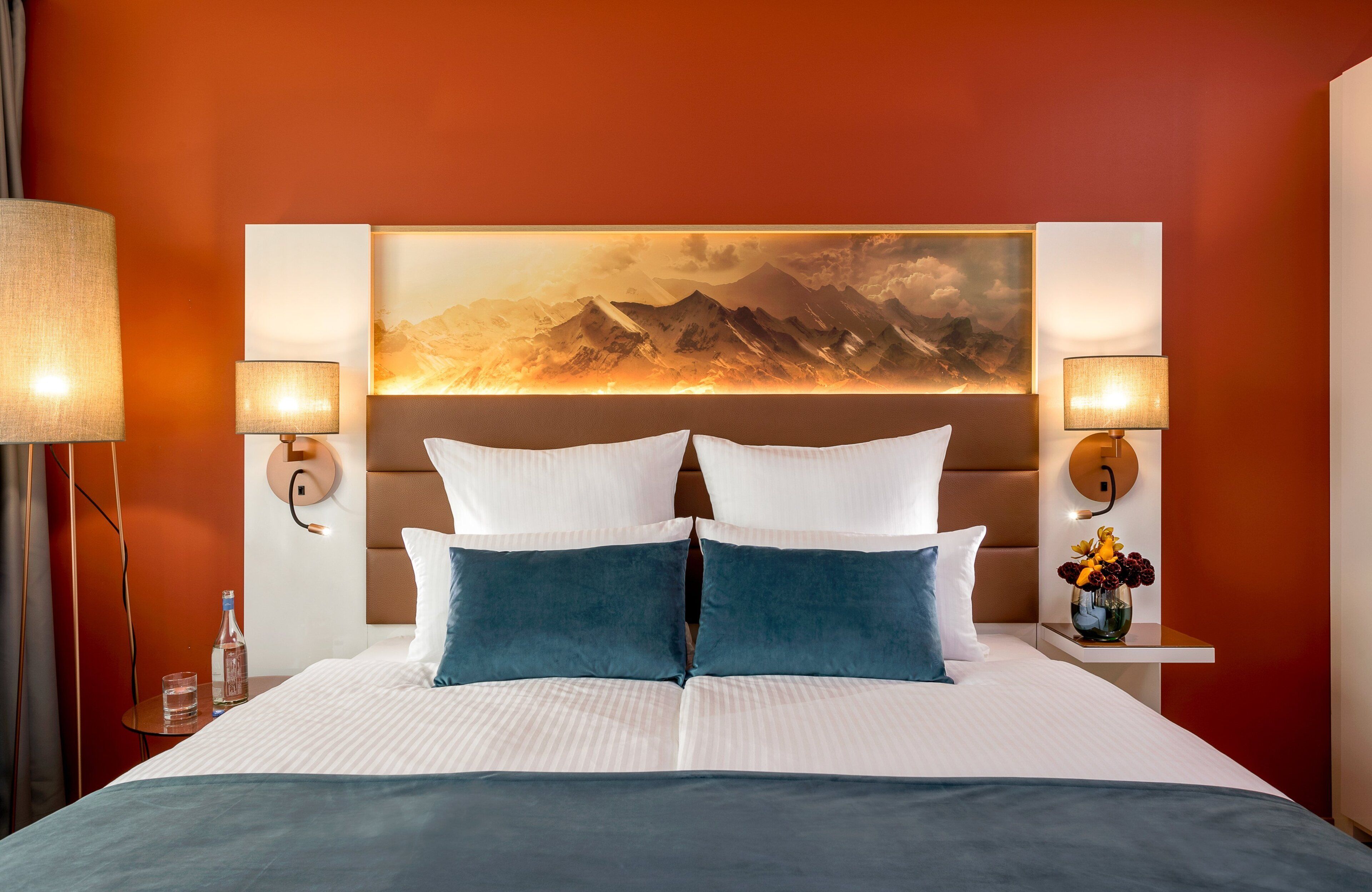 A hotel room with a large bed, two wall-mounted lamps, a painting of mountains, and a small table with a vase of flowers.