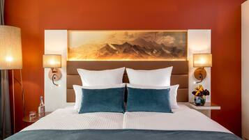 A hotel room with a large bed, two wall-mounted lamps, a painting of mountains, and a small table with a vase of flowers.