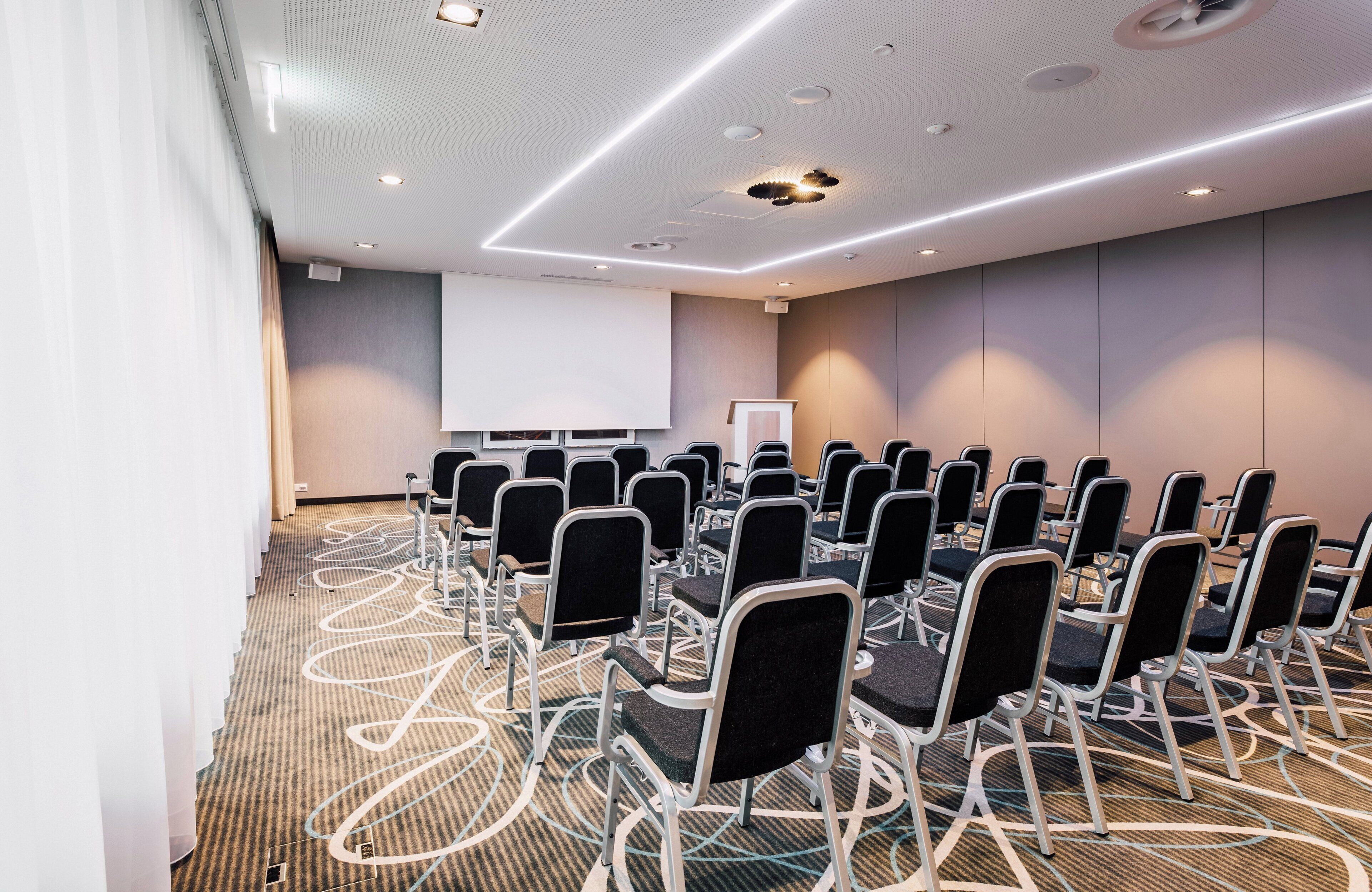 Un salón de conferencias con filas de sillas, una pantalla de proyector y piso alfombrado.