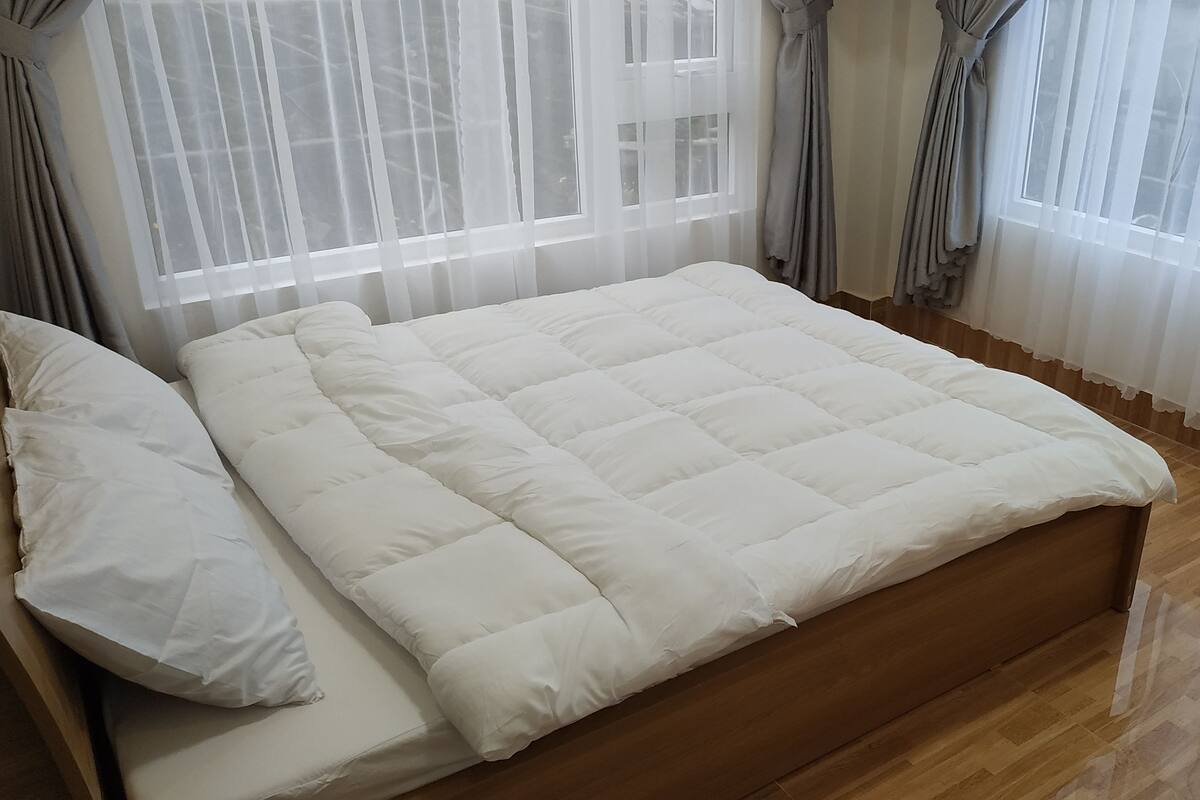 A neatly made bed with a white duvet and pillows, a wooden headboard, and a window with sheer curtains.