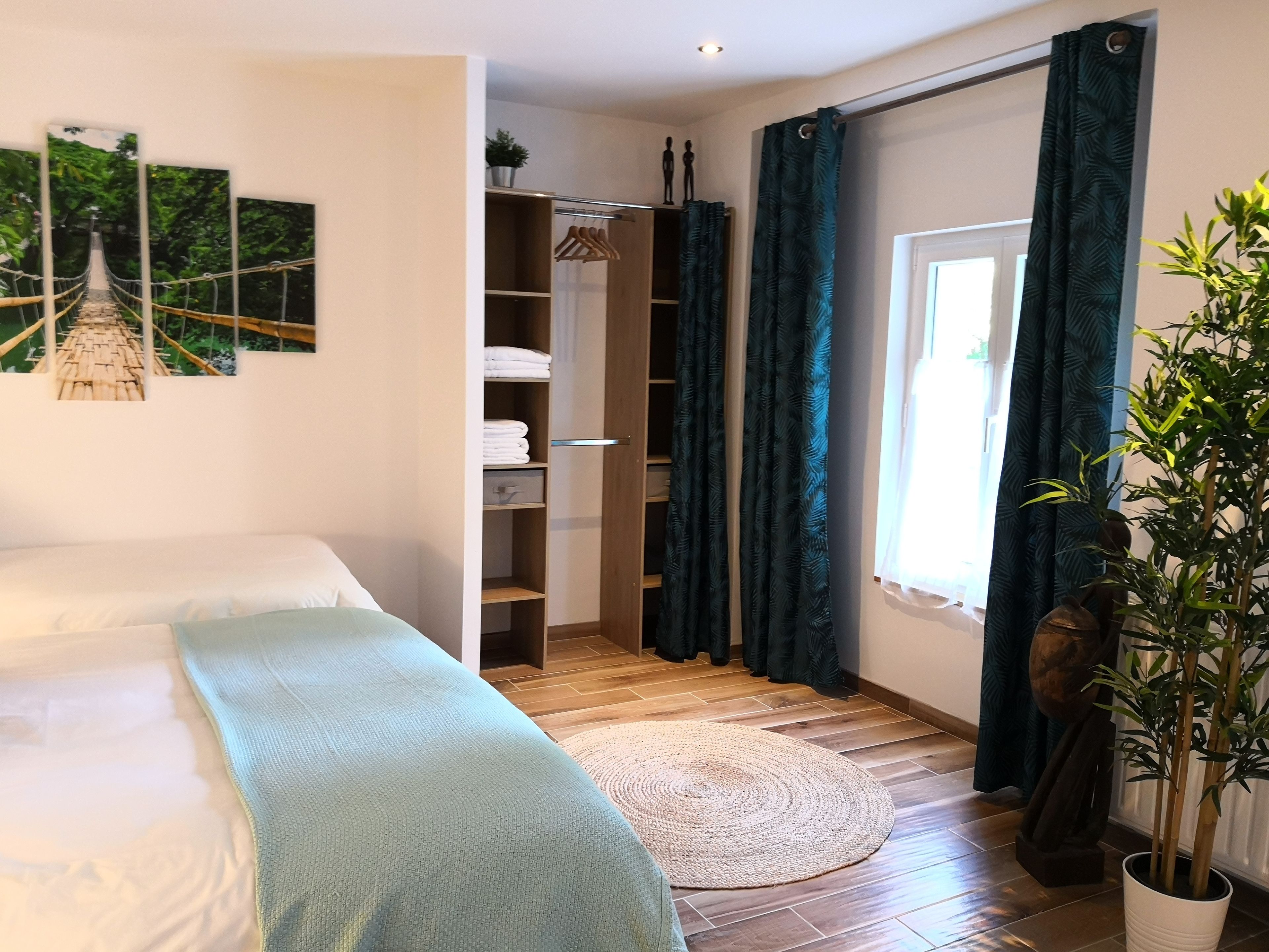 Ein Schlafzimmer mit Bett, Kleiderschrank, einem kleinen Fenster mit Vorhängen und einer Topfpflanze.