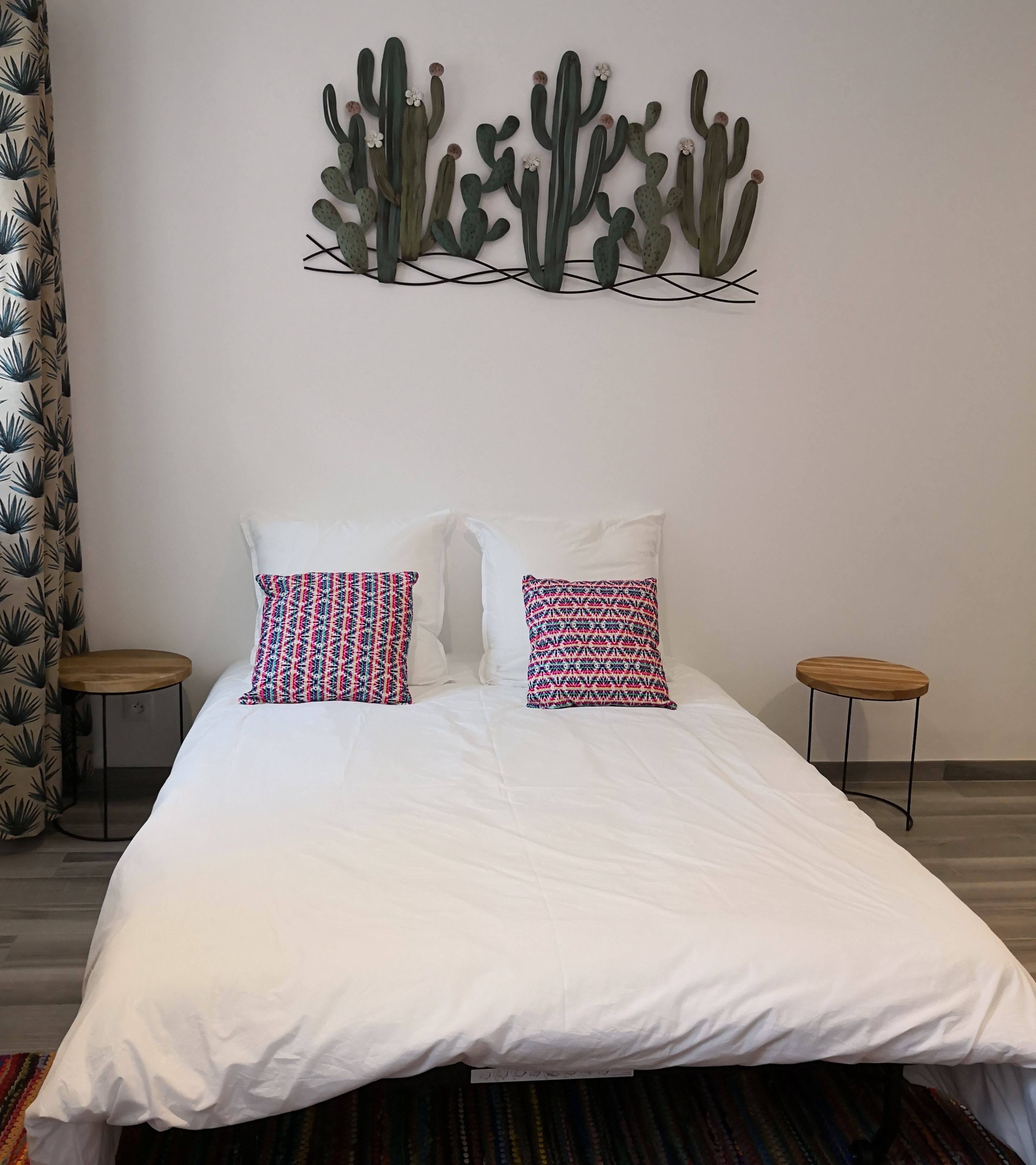 Una cama con ropa de cama blanca, dos almohadas estampadas y un cuadro de pared con un cactus.