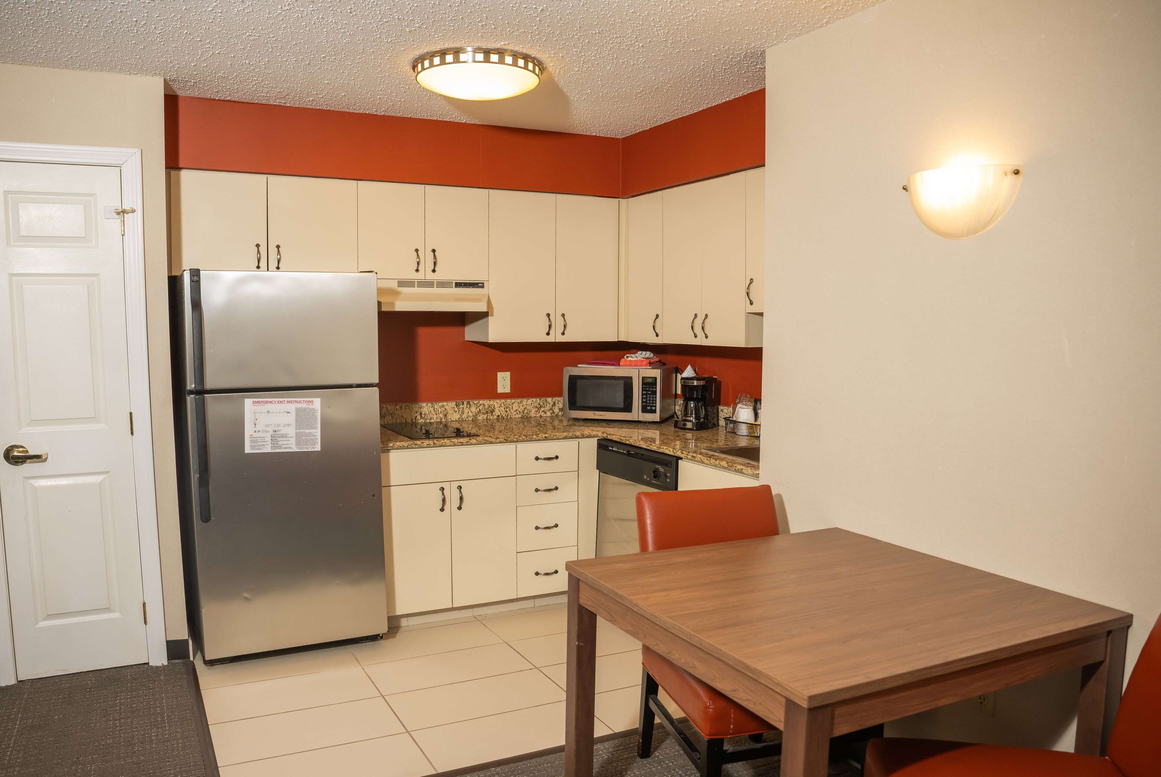 A compact kitchen with white cabinets, a stainless steel refrigerator, a microwave, and a coffee maker. There is a wooden table with two chairs.