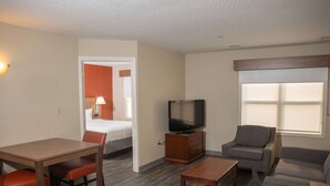 A hotel room with a wooden dining table, two chairs, a sofa, a TV on a wooden cabinet, and a window with blinds.