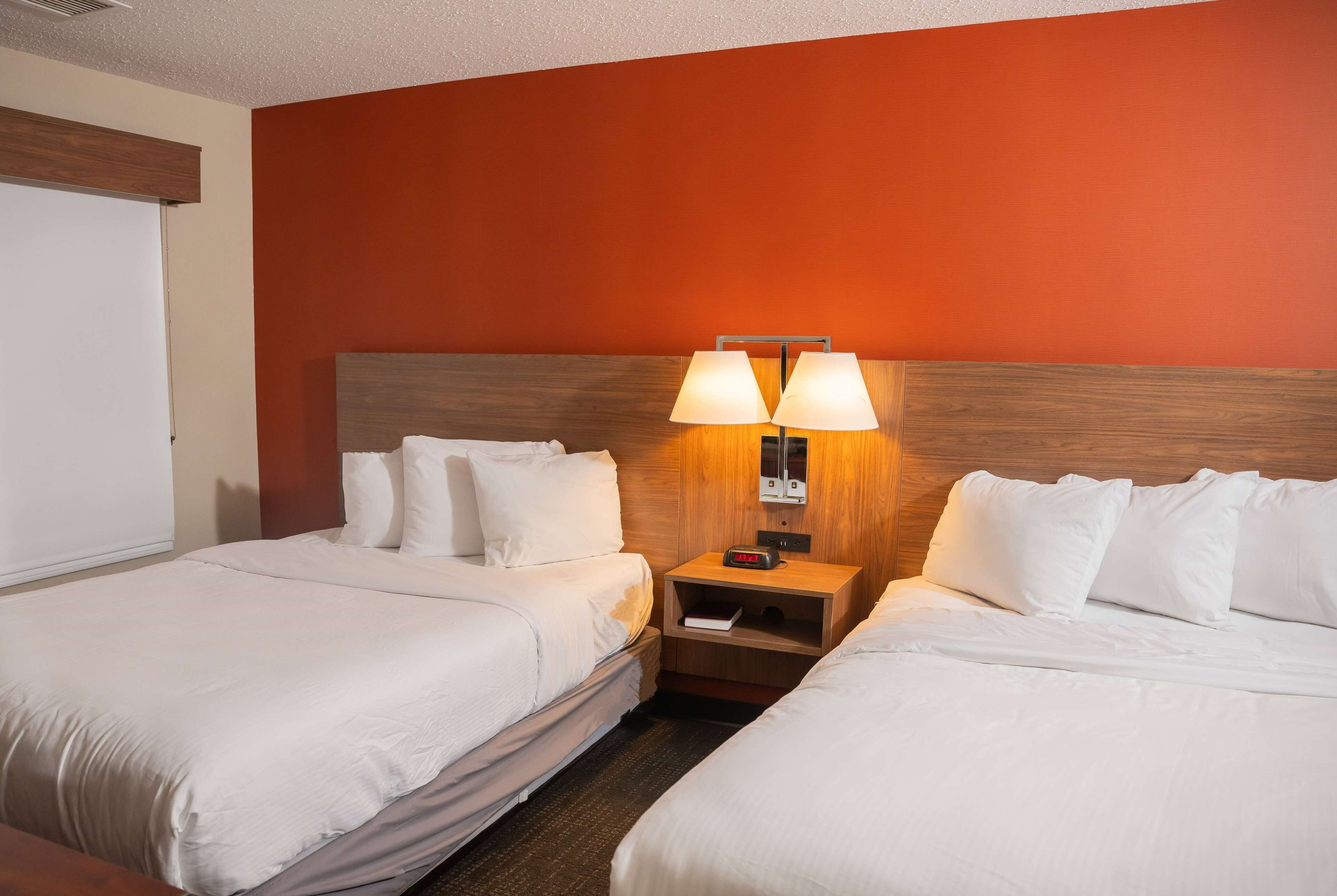 A hotel room with two beds, a wooden headboard, a nightstand with a lamp, and a white door.