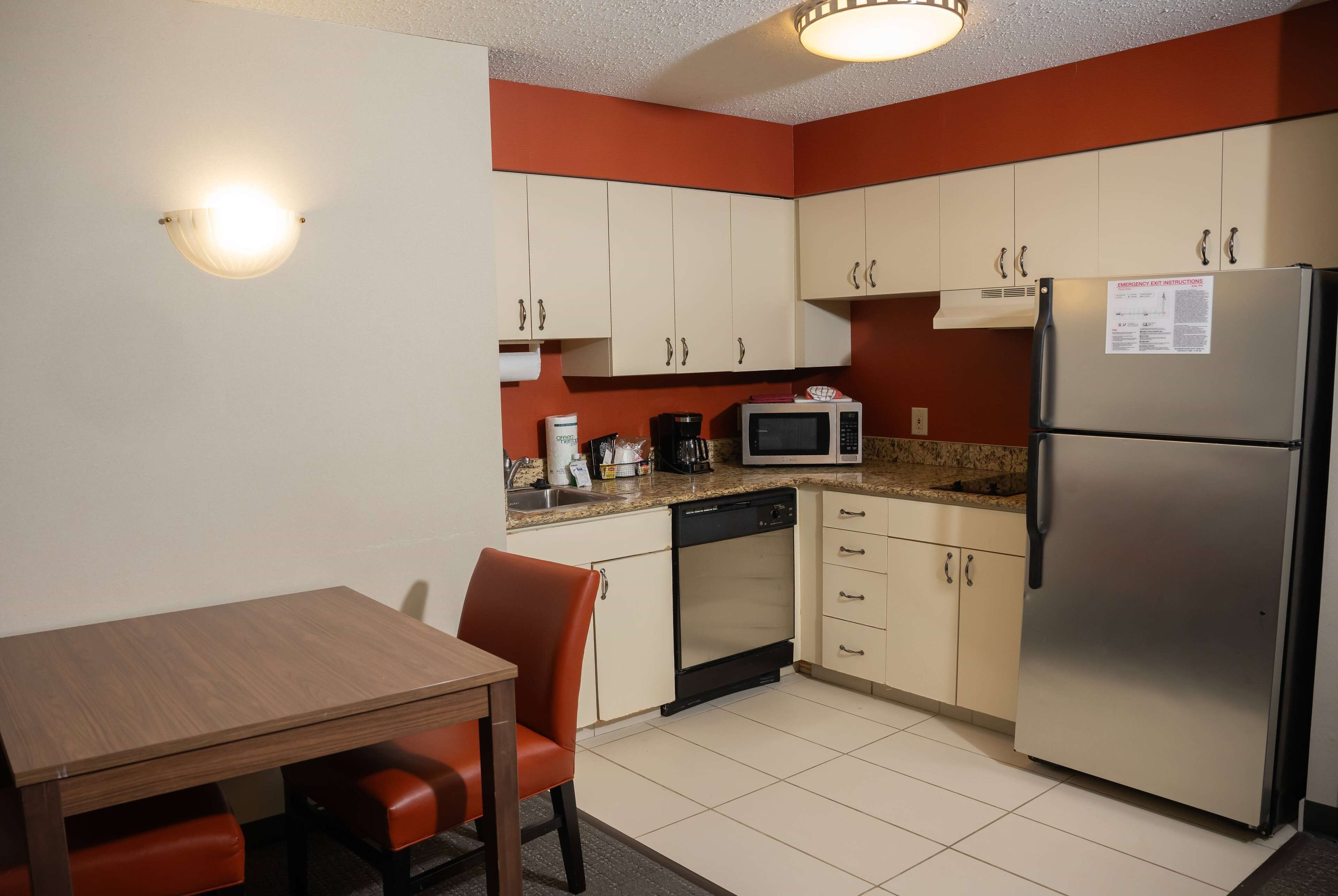 A compact kitchen with white cabinets, a stainless steel refrigerator, a microwave, and a dishwasher. There is a wooden table with two chairs.