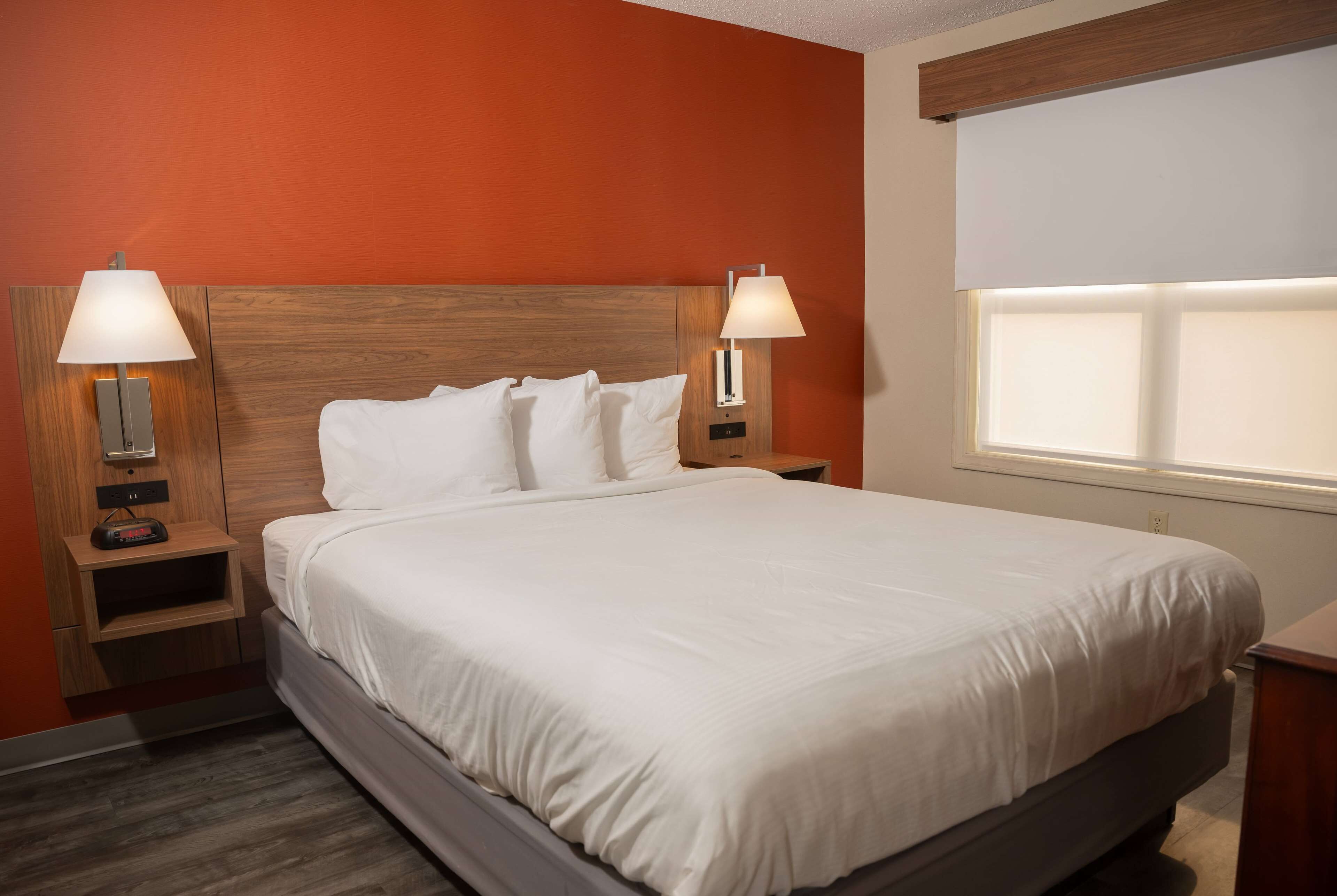 A neatly made bed in a hotel room with a wooden headboard, two bedside lamps, a nightstand with a clock, and a window with blinds.