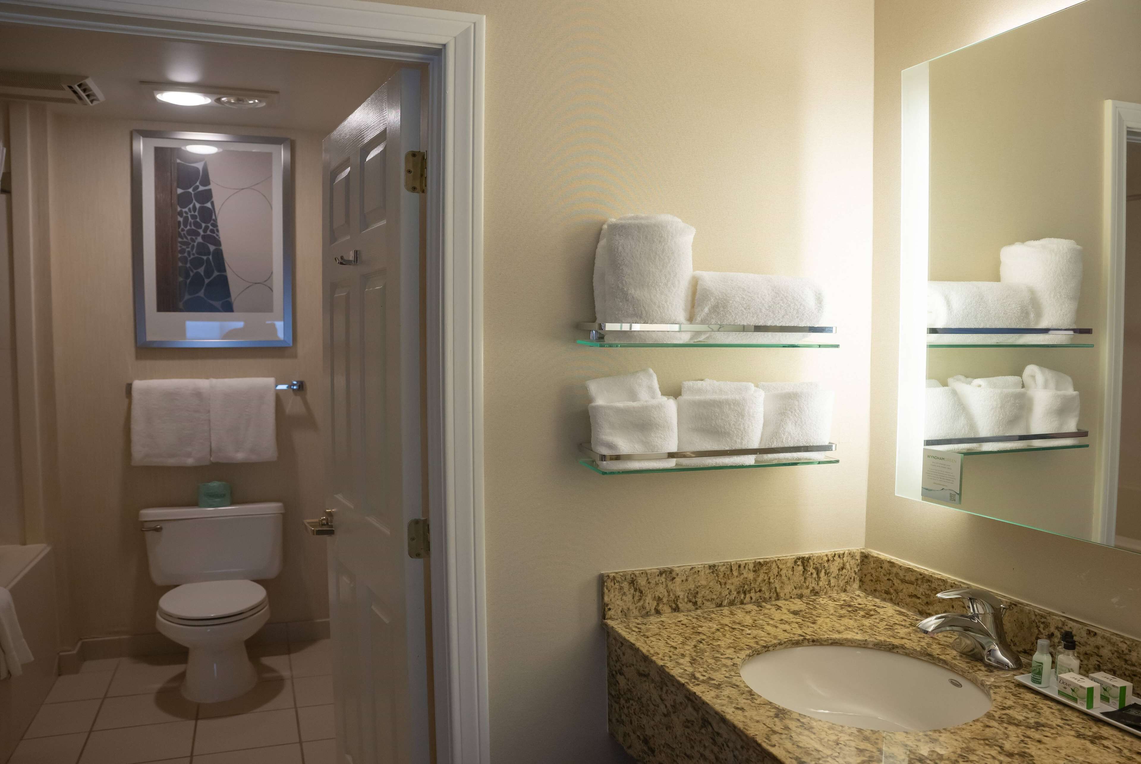 A bathroom with a toilet, a sink with a granite countertop, and glass shelves holding white towels.