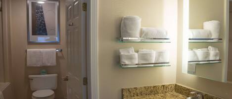 A bathroom with a toilet, a sink with a granite countertop, and glass shelves holding white towels.