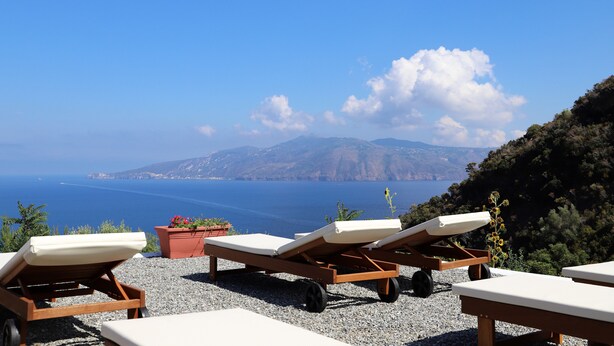 Terraza frente al mar con sillones de descanso y plantas en macetas, con vistas a un paisaje costero con montañas y el océano.