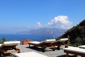 Une terrasse en bord de mer, dotée de chaises longues et de plantes en pot, qui offre une vue sur un paysage côtier avec des montagnes et la mer.