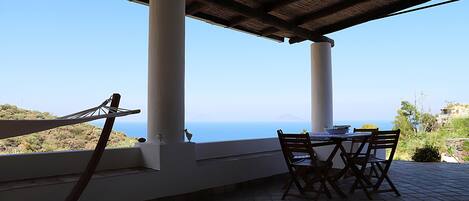 A terrace with a table and chairs, overlooking a scenic view of the sea and hills.