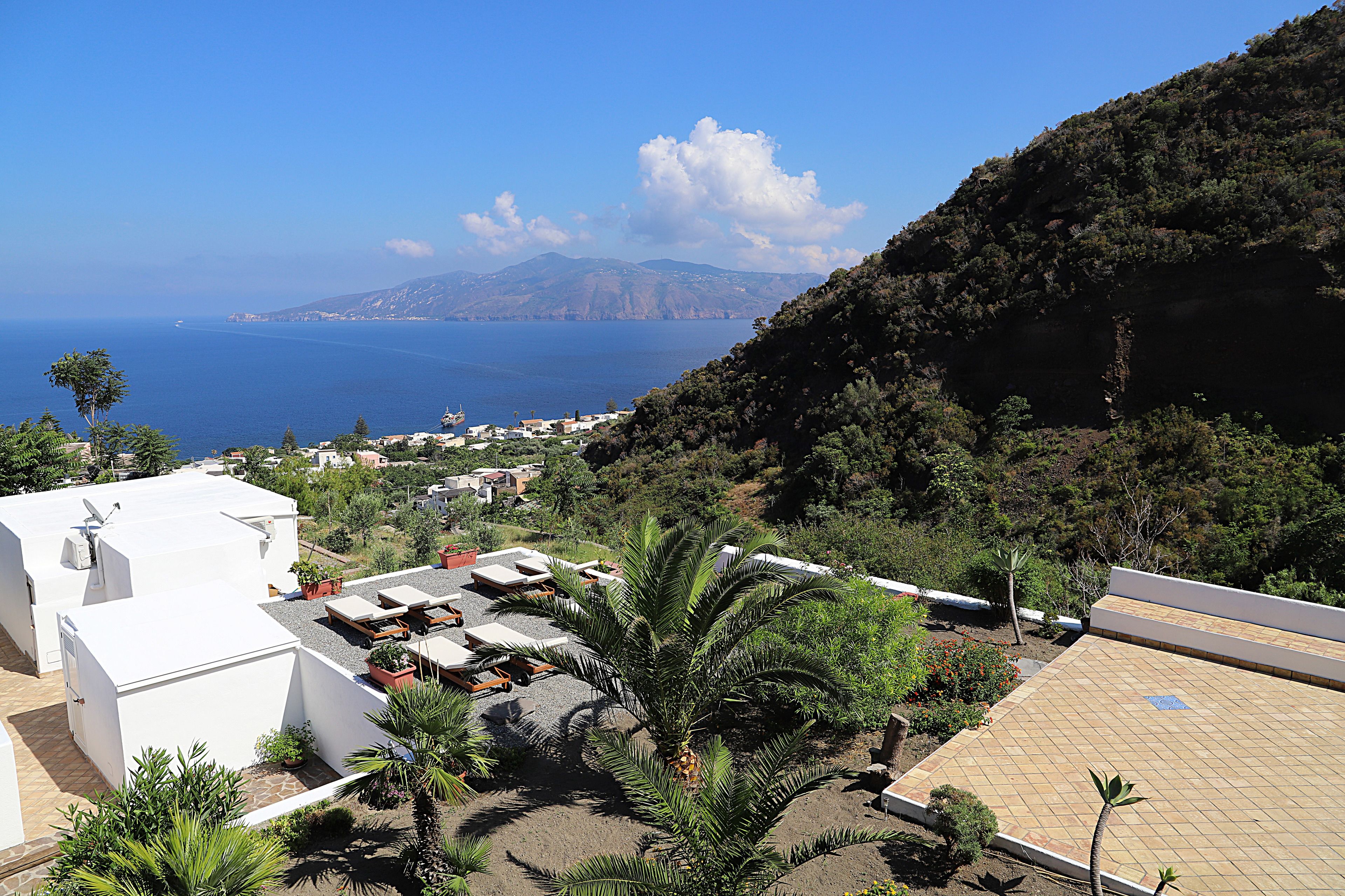 A rooftop terrace with outdoor seating, a palm tree, and a view of the sea and distant mountains.