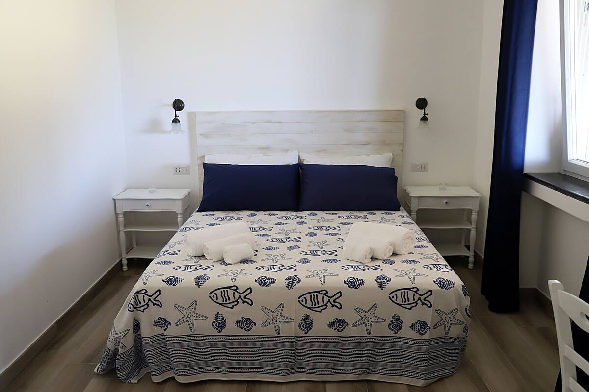 Un lit bien fait, recouvert d’une courtepointe à motifs d’étoiles de mer et de poissons, avec deux oreillers et deux tables de chevet dotées de lampes.