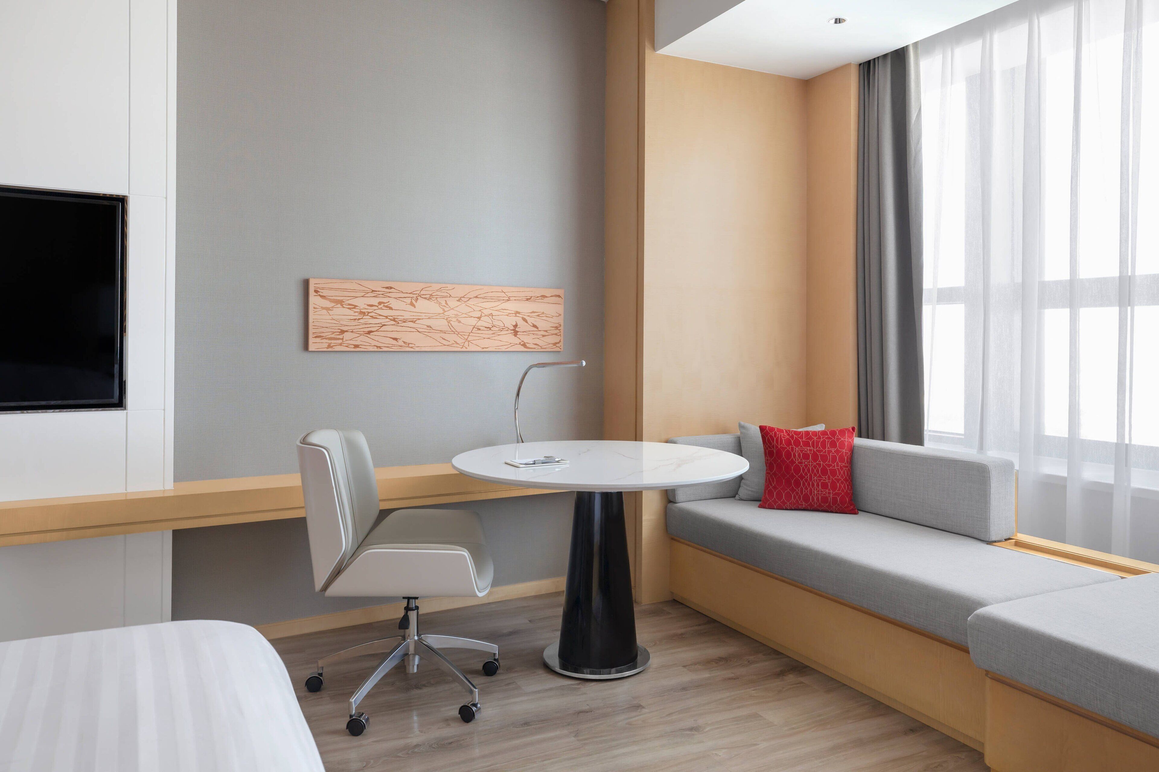 A modern hotel room with a sofa, a small round table, a chair, and a flat-screen TV mounted on the wall.
