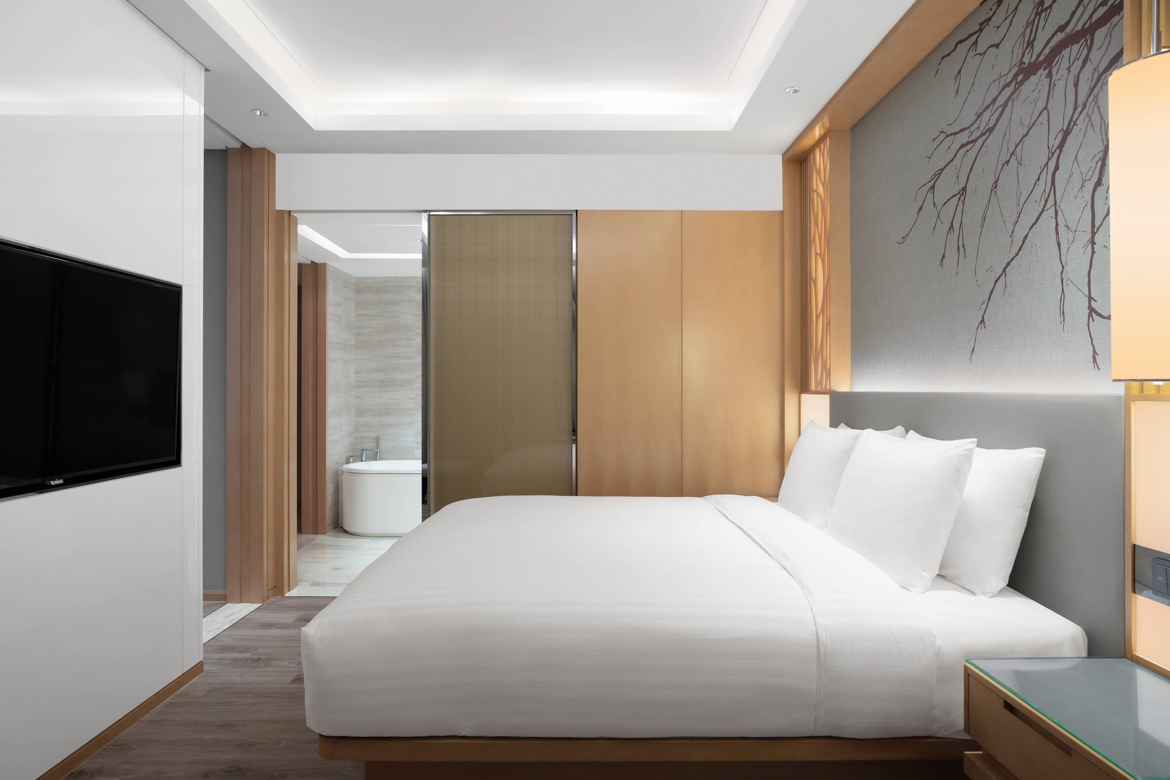 A modern hotel room with a large bed, a flat-screen TV, a wooden headboard, and a large artwork on the wall.