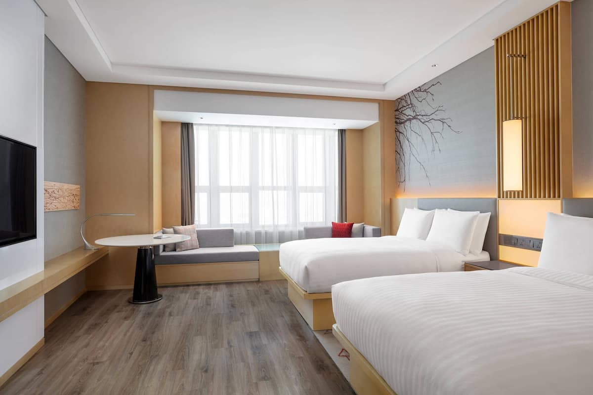 A hotel room with two beds, a seating area with a table, a large window with sheer curtains, a wall-mounted TV, and a modern lamp.