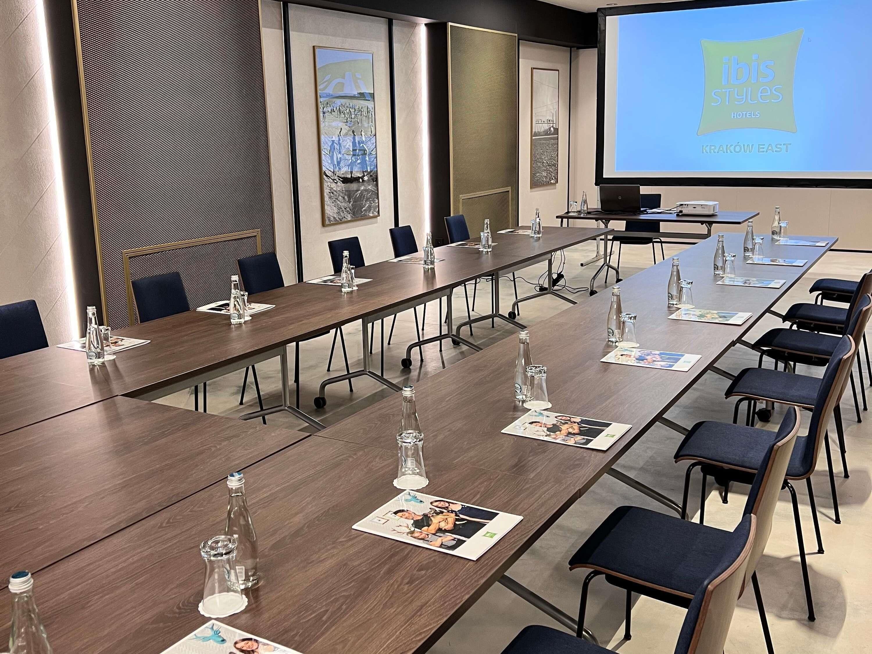 A conference room with long tables, chairs, water bottles, and a presentation screen displaying 'ibis Styles Hotels Krakow East'.