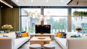 Un salon moderne doté d’un grand téléviseur à écran plat, de deux canapés ornés de coussins colorés, d’une table basse en bois et d’une grande fenêtre offrant une vue sur les arbres et les bâtiments.