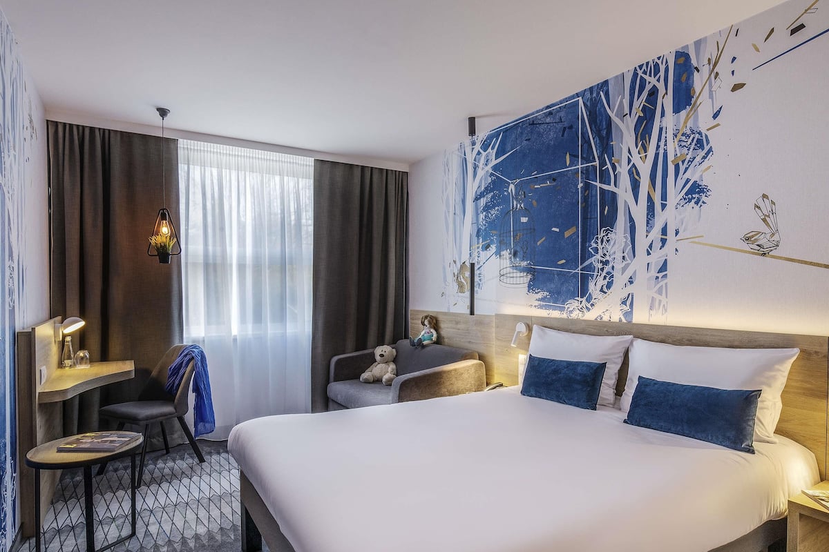 Een hotelkamer met een groot bed, een bureau met een lamp, een stoel, een teddybeer en een groot abstract schilderij aan de muur.